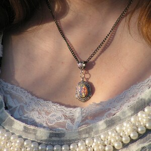 May include: A silver chain necklace with an oval-shaped pendant featuring an iridescent opal stone. The necklace is worn by a person with long brown hair and a white lace top.