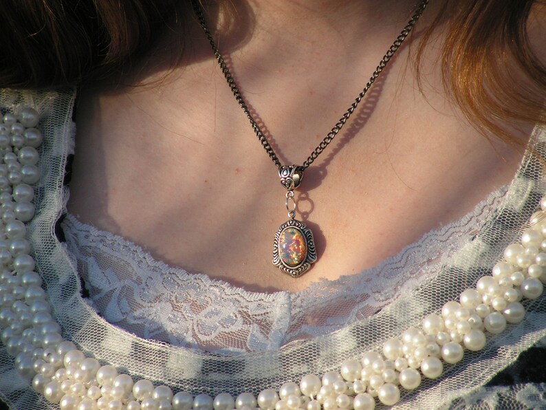 May include: A silver chain necklace with an oval-shaped pendant featuring an iridescent opal stone. The necklace is worn by a person with long brown hair and a white lace top.