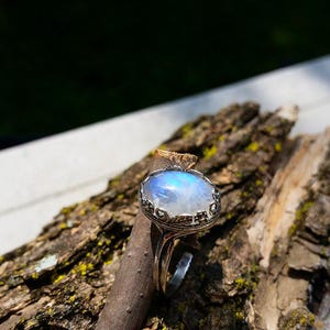 May include: A silver ring with a large, oval, iridescent moonstone. The stone has a blue and white glow. The ring is set on a piece of wood with a rough bark texture, with a blurred green background.