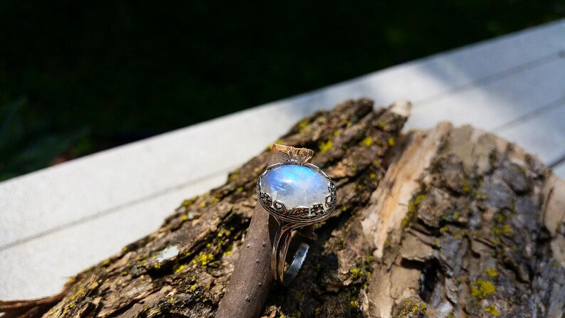 May include: A silver ring with a large, oval, iridescent moonstone. The stone has a blue and white glow. The ring is set on a piece of wood with a rough bark texture, with a blurred green background.