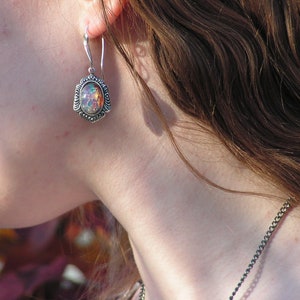 May include: A silver earring with an iridescent opal stone set in a decorative frame. The earring is worn by a person with long brown hair.