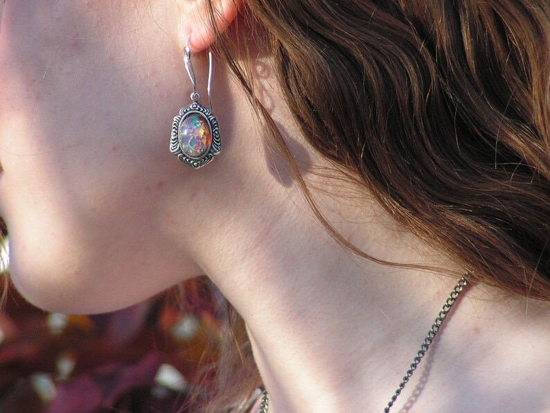 May include: A silver earring with an iridescent opal stone set in a decorative frame. The earring is worn by a person with long brown hair.