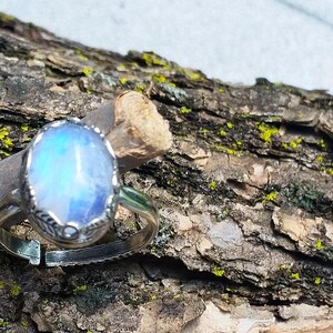 May include: A silver ring with an oval, iridescent, blue gemstone. The ring is set on a piece of wood with a rough, textured bark. The gemstone has a pearlescent, glowing appearance, and the ring's band is adjustable.