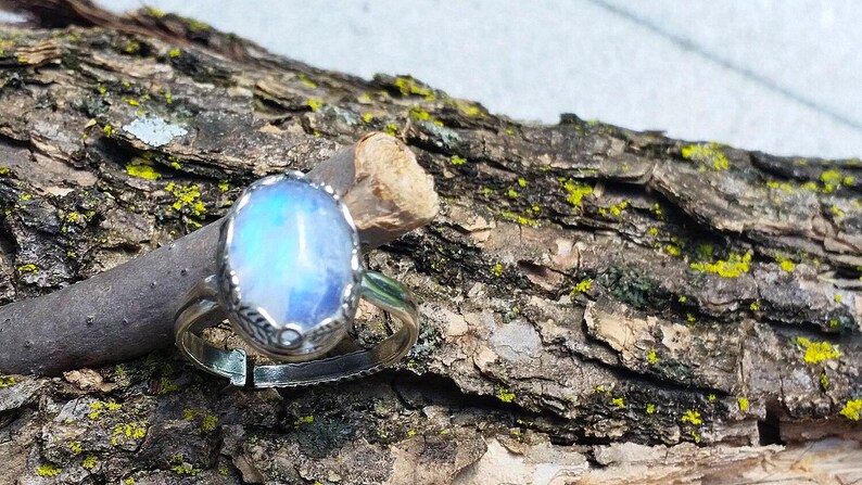 May include: A silver ring with an oval, iridescent, blue gemstone. The ring is set on a piece of wood with a rough, textured bark. The gemstone has a pearlescent, glowing appearance, and the ring's band is adjustable.