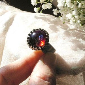 May include: A silver ring with a dark red and blue iridescent gemstone set in a silver bezel. The ring is on a finger and the background is a white fabric.