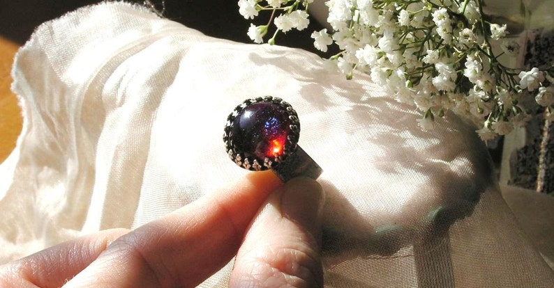 May include: A silver ring with a dark red and blue iridescent gemstone set in a silver bezel. The ring is on a finger and the background is a white fabric.