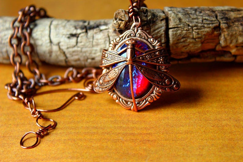 K&ouml;nnte beinhalten: Eine kupferfarbene Libellen-Anh&auml;nger-Halskette. Der Anh&auml;nger zeigt ein Libellen-Design mit detaillierten Fl&uuml;geln und einem zentralen Stein mit roten und blauen Farbt&ouml;nen. Die Halskette ist ebenfalls kupferfarben und liegt auf einem Holzscheit.