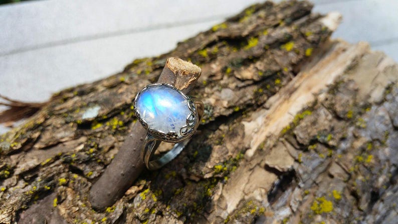 May include: A silver ring with a large, oval, iridescent moonstone. The stone has a blue and white glow. The ring is set on a piece of textured wood.