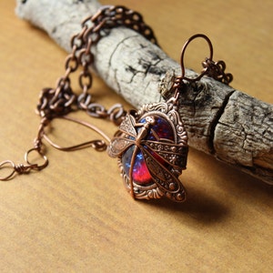 K&ouml;nnte beinhalten: Kupferfarbene Libellen-Anh&auml;nger-Halskette. Der Anh&auml;nger zeigt ein detailliertes Libellen-Design &uuml;ber einem leuchtenden rot-blauen Stein. Die Halskette wird auf einem St&uuml;ck verwittertem Holz pr&auml;sentiert, das die filigrane Metallarbeit und die Farbe des Steins zeigt.