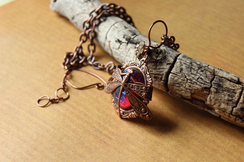 K&ouml;nnte beinhalten: Kupferfarbene Libellen-Anh&auml;nger-Halskette. Der Anh&auml;nger zeigt ein detailliertes Libellen-Design &uuml;ber einem leuchtenden rot-blauen Stein. Die Halskette wird auf einem St&uuml;ck verwittertem Holz pr&auml;sentiert, das die filigrane Metallarbeit und die Farbe des Steins zeigt.