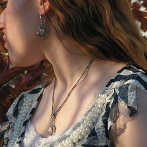 May include: A woman wearing a white and black lace top with a silver necklace and earrings. The necklace has a small pendant with a floral design.