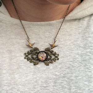 Puede incluir: Un collar de estilo vintage con una cadena y colgante de color bronce. El colgante presenta una piedra central, iridiscente y circular, rodeada por un marco ornamental de inspiraci&oacute;n floral. Dos peque&ntilde;os p&aacute;jaros est&aacute;n colocados encima.