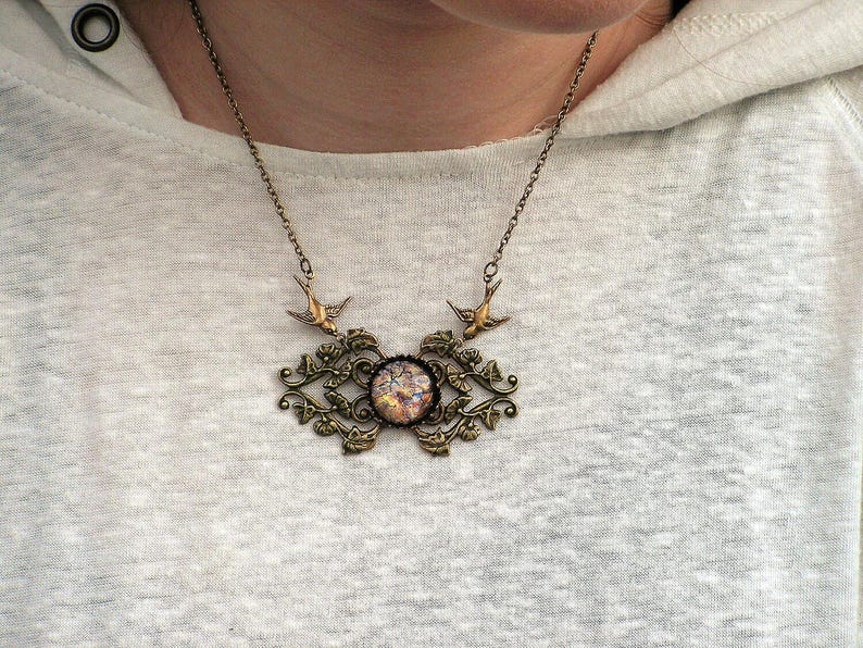 Puede incluir: Un collar de estilo vintage con una cadena y colgante de color bronce. El colgante presenta una piedra central, iridiscente y circular, rodeada por un marco ornamental de inspiraci&oacute;n floral. Dos peque&ntilde;os p&aacute;jaros est&aacute;n colocados encima.