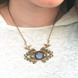 Puede incluir: Un collar de estilo vintage con una cadena y colgante de color bronce. El colgante presenta una piedra iridiscente central rodeada de filigranas ornamentadas y con hojas y dos peque&ntilde;os abalorios de p&aacute;jaros. El collar se lleva sobre una camiseta blanca.