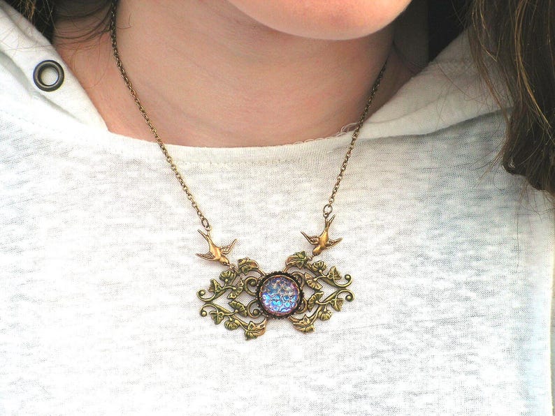 Puede incluir: Un collar de estilo vintage con una cadena y colgante de color bronce. El colgante presenta una piedra iridiscente central rodeada de filigranas ornamentadas y con hojas y dos peque&ntilde;os abalorios de p&aacute;jaros. El collar se lleva sobre una camiseta blanca.