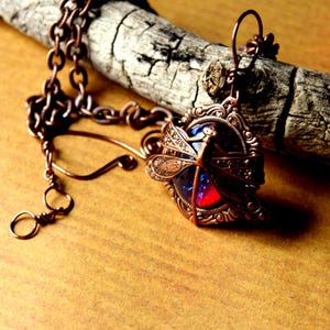 K&ouml;nnte beinhalten: Eine kupferfarbene Libellen-Anh&auml;nger-Halskette mit detailliertem Filigran-Design. Der Anh&auml;nger hat ein blaues und rotes Glaszentrum. Die Halskette wird auf einem St&uuml;ck verwittertem Holz pr&auml;sentiert.