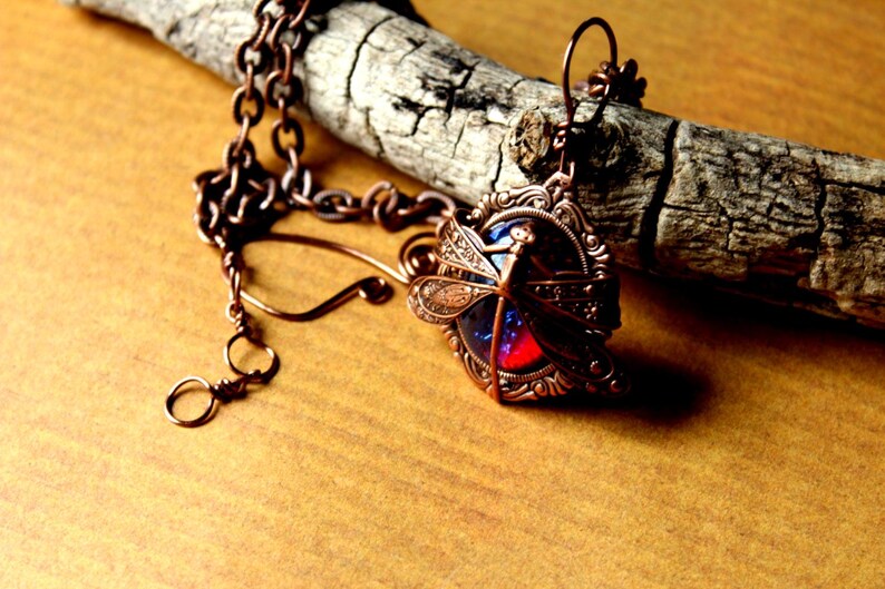 K&ouml;nnte beinhalten: Eine kupferfarbene Libellen-Anh&auml;nger-Halskette mit detailliertem Filigran-Design. Der Anh&auml;nger hat ein blaues und rotes Glaszentrum. Die Halskette wird auf einem St&uuml;ck verwittertem Holz pr&auml;sentiert.