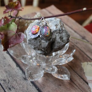 May include: A pair of silver earrings with iridescent opal stones set in ornate silver frames. The earrings are displayed on a clear glass lotus flower base and a grey rock.