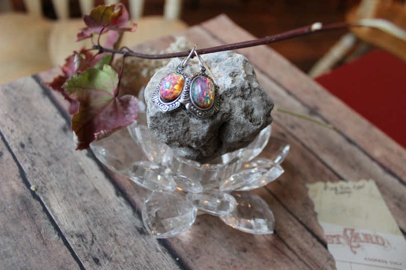 May include: A pair of silver earrings with iridescent opal stones set in ornate silver frames. The earrings are displayed on a clear glass lotus flower base and a grey rock.