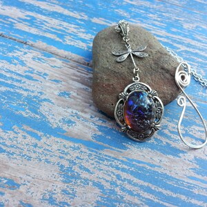 May include: A silver necklace with a blue opal pendant and a dragonfly charm. The necklace is on a rustic blue wooden background.