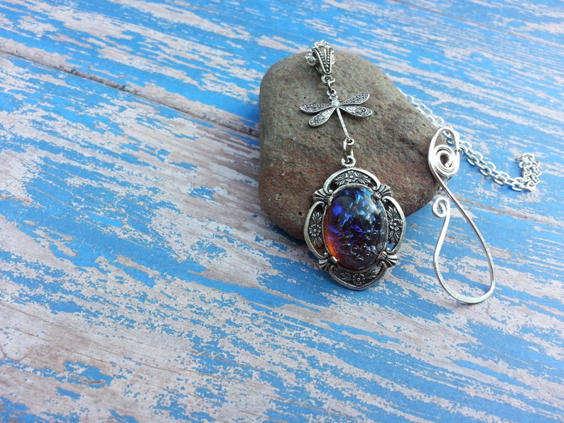 May include: A silver necklace with a blue opal pendant and a dragonfly charm. The necklace is on a rustic blue wooden background.