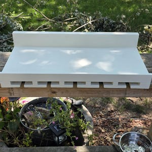 May include: A white wooden wall-mounted shelf featuring a flat top surface with a back ledge and a bottom section designed with multiple T-shaped cutouts for hanging stemware glasses, providing a functional storage solution for kitchen or bar areas.