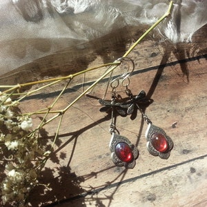 May include: A pair of silver dangle earrings with red gemstone accents. The earrings feature a dragonfly design and are hanging from a wooden surface.
