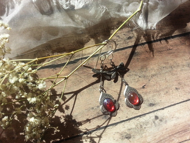 May include: A pair of silver dangle earrings with red gemstone accents. The earrings feature a dragonfly design and are hanging from a wooden surface.