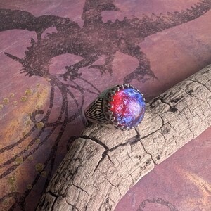 May include: A silver ring with a large, round, blue and red opal gemstone. The ring has a detailed, geometric design on the band.