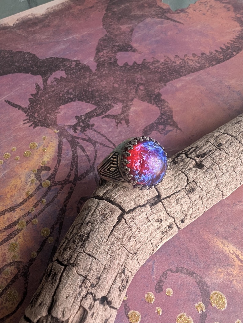 May include: A silver ring with a large, round, blue and red opal gemstone. The ring has a detailed, geometric design on the band.
