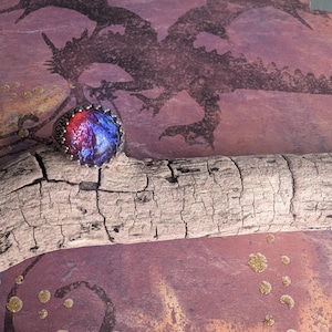 May include: A silver ring with a blue and red opal stone set in a bezel setting. The ring is on a piece of driftwood in front of a background with a dragon silhouette and gold glitter.
