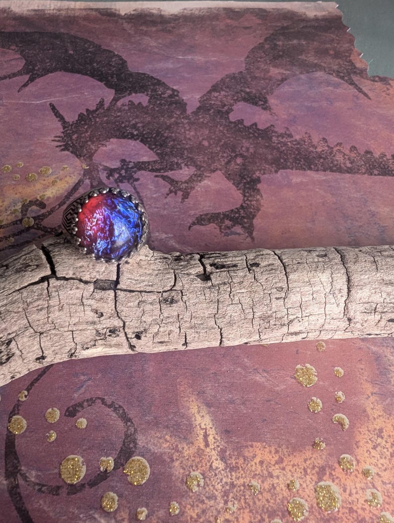 May include: A silver ring with a blue and red opal stone set in a bezel setting. The ring is on a piece of driftwood in front of a background with a dragon silhouette and gold glitter.