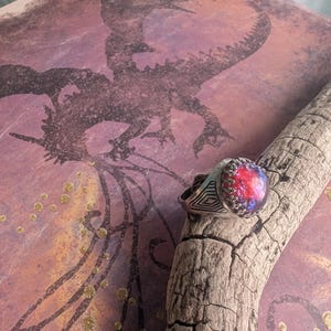 May include: A silver ring with a large, round, iridescent gemstone set in the center. The gemstone has shades of red, blue, and purple. The ring is on a piece of driftwood with a background of a dragon silhouette.