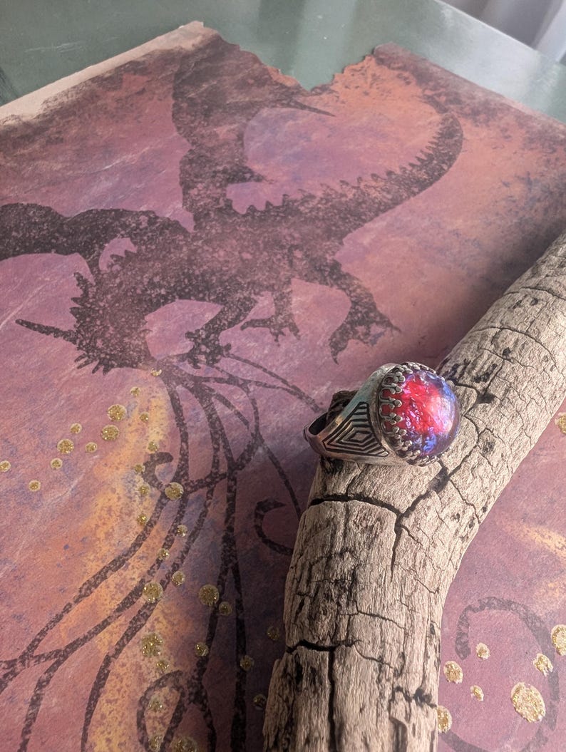 May include: A silver ring with a large, round, iridescent gemstone set in the center. The gemstone has shades of red, blue, and purple. The ring is on a piece of driftwood with a background of a dragon silhouette.