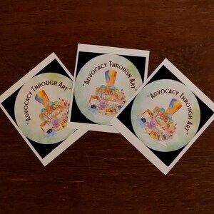 May include: Three white square stickers with a black background and a colourful illustration of a paintbrush with the text "Advocacy Through Art" in a circle.