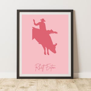 Puede incluir: Una silueta rosa de un vaquero montando un toro con el texto "Rhett Eaton" debajo. La imagen está enmarcada en negro.
