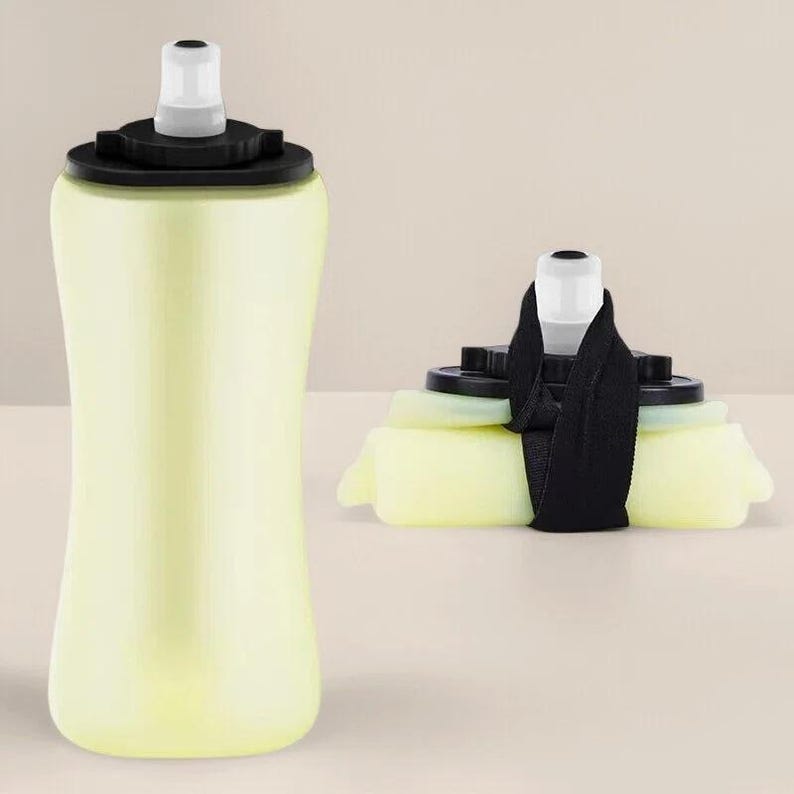 Puede incluir: Una botella de agua plegable de color amarillo claro con una tapa negra y una boquilla blanca. La botella se muestra tanto llena como plegada, con una correa negra que asegura la versi&oacute;n plegada. Ideal para actividades al aire libre.