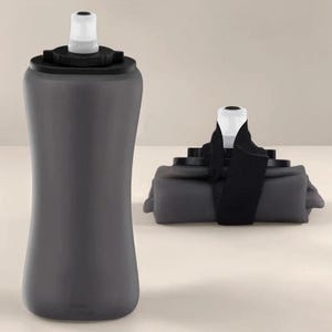 Puede incluir: Una botella de agua plegable de color gris oscuro con una boquilla blanca y una tapa negra. La botella se muestra tanto en posici&oacute;n vertical como plegada, con una correa negra que asegura la versi&oacute;n plegada. La botella parece estar hecha de un material flexible.