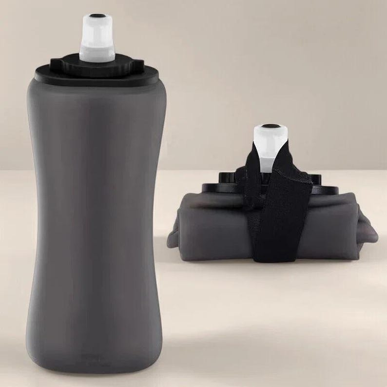 Puede incluir: Una botella de agua plegable de color gris oscuro con una boquilla blanca y una tapa negra. La botella se muestra tanto en posici&oacute;n vertical como plegada, con una correa negra que asegura la versi&oacute;n plegada. La botella parece estar hecha de un material flexible.