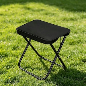 Könnte beinhalten: Ein schwarzer Klapphocker mit gepolsterter Sitzfläche. Der Hocker hat ein schwarzes Metallgestell und steht auf einer grünen Wiese. Die Sitzfläche ist rechteckig und scheint aus einem weichen, gepolsterten Material zu bestehen.