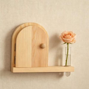 May include: A light-coloured wooden wall shelf featuring an arched door design with a small round knob. A single peach-coloured rose is placed inside a clear, cylindrical glass test tube vase that is attached to the side of the wooden shelf.