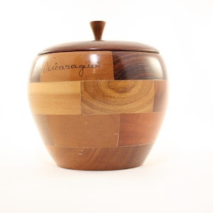 May include: A wooden apple-shaped container with a lid and a small handle. The container is made of various wood tones, with the word "Nicaragua" written on the side. The container is on a white background.