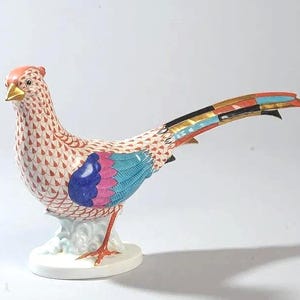 May include: A decorative porcelain pheasant figurine. The bird features a detailed design with a red, white, and blue pattern on its body, with accents of pink, blue, and gold. The pheasant stands on a white base.