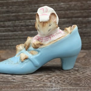 May include: A blue ceramic shoe with a white and pink mouse figurine sitting inside. The mouse is wearing a pink and white dress and a white hat.