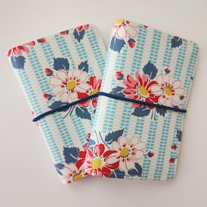 May include: Two fabric-covered journals with a floral pattern. The covers feature a white background with light blue vertical stripes and a grid pattern. Large red and white flowers with yellow centers and blue leaves are printed on the covers. A dark blue elastic band secures the journals.