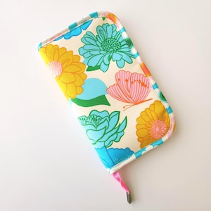 May include: A rectangular, cream-colored case with a colorful floral and butterfly pattern. The design features yellow, blue, and pink flowers, and a pink butterfly. The edges are trimmed with a blue and white striped fabric. A pink ribbon is attached.