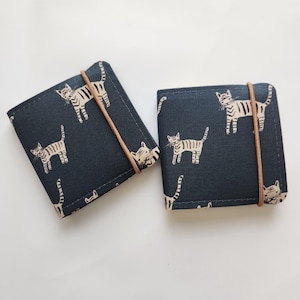 May include: Two square fabric wallets in a dark navy blue color featuring a repeating pattern of cream-colored tabby cats. Each wallet is secured with a thin, vertical tan elastic band that wraps around the center of the folded fabric exterior.