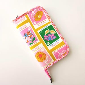 May include: A rectangular, zippered pouch with a floral seed packet design. The pouch features a pink and yellow color scheme with text that reads "Seeds of Free Time" and "Seeds of Inspiration". The edges are trimmed with a pink and white checkered pattern.