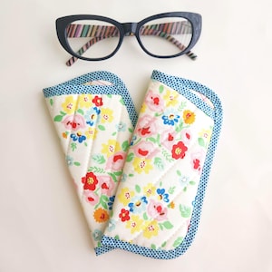 May include: A pair of dark blue cat-eye glasses with a multicolored striped inner frame, positioned above a quilted fabric eyeglass case featuring a white background with a vibrant floral pattern in red, pink, yellow, and blue, finished with blue polka dot trim.