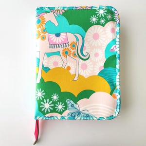 May include: A fabric-covered journal featuring a whimsical design. The cover showcases a pink unicorn adorned with orange flowers, set against a backdrop of green, yellow, and pink clouds. A blue butterfly and white daisies complete the design. A pink ribbon bookmark is attached.